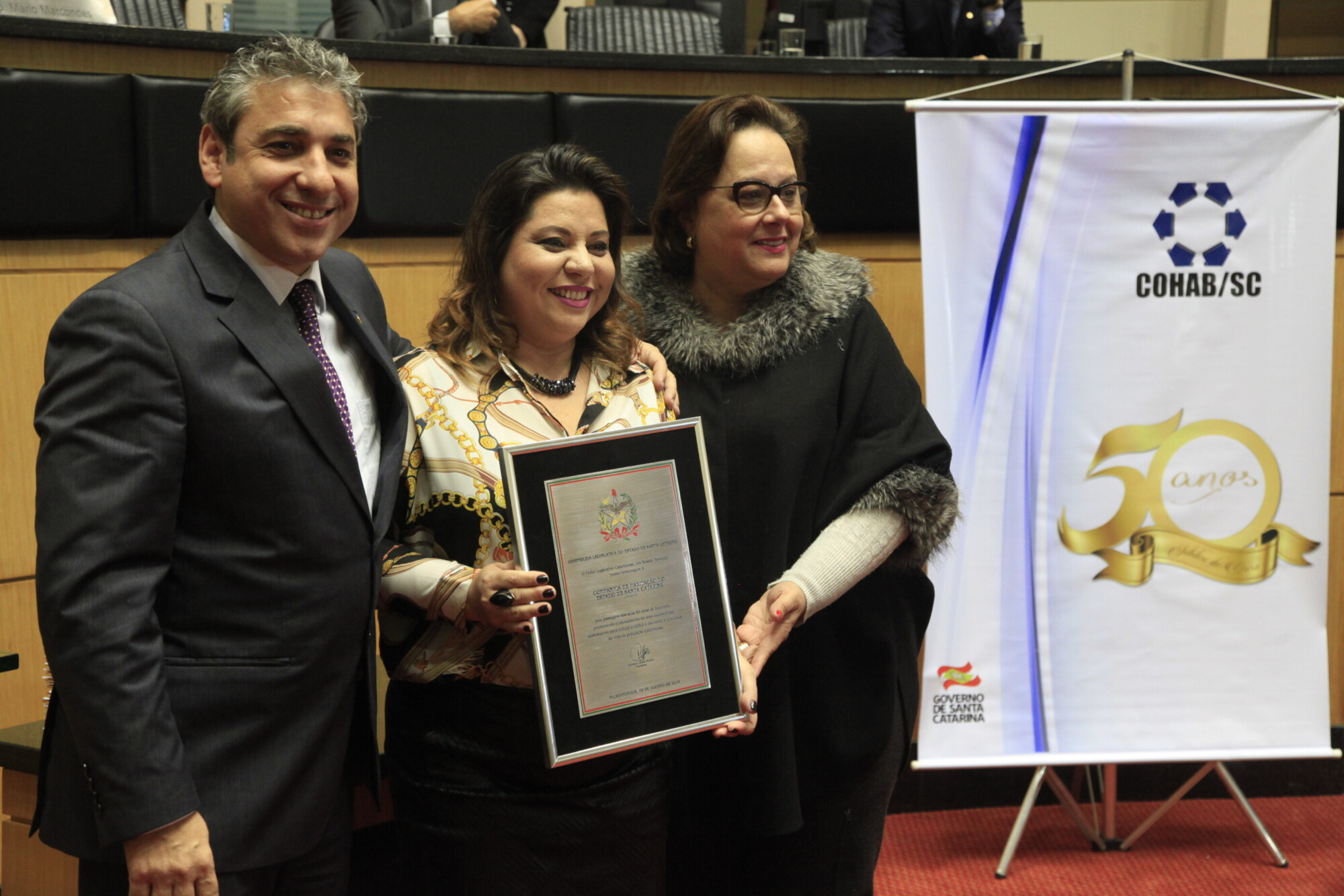 Deputados Mário Marcondes (PSDB) e Dirce Heiderscheidt (PMDB) entregam placa à diretora-presidente da Cohab/SC, Alessandra de Andrade Klettenberg.
