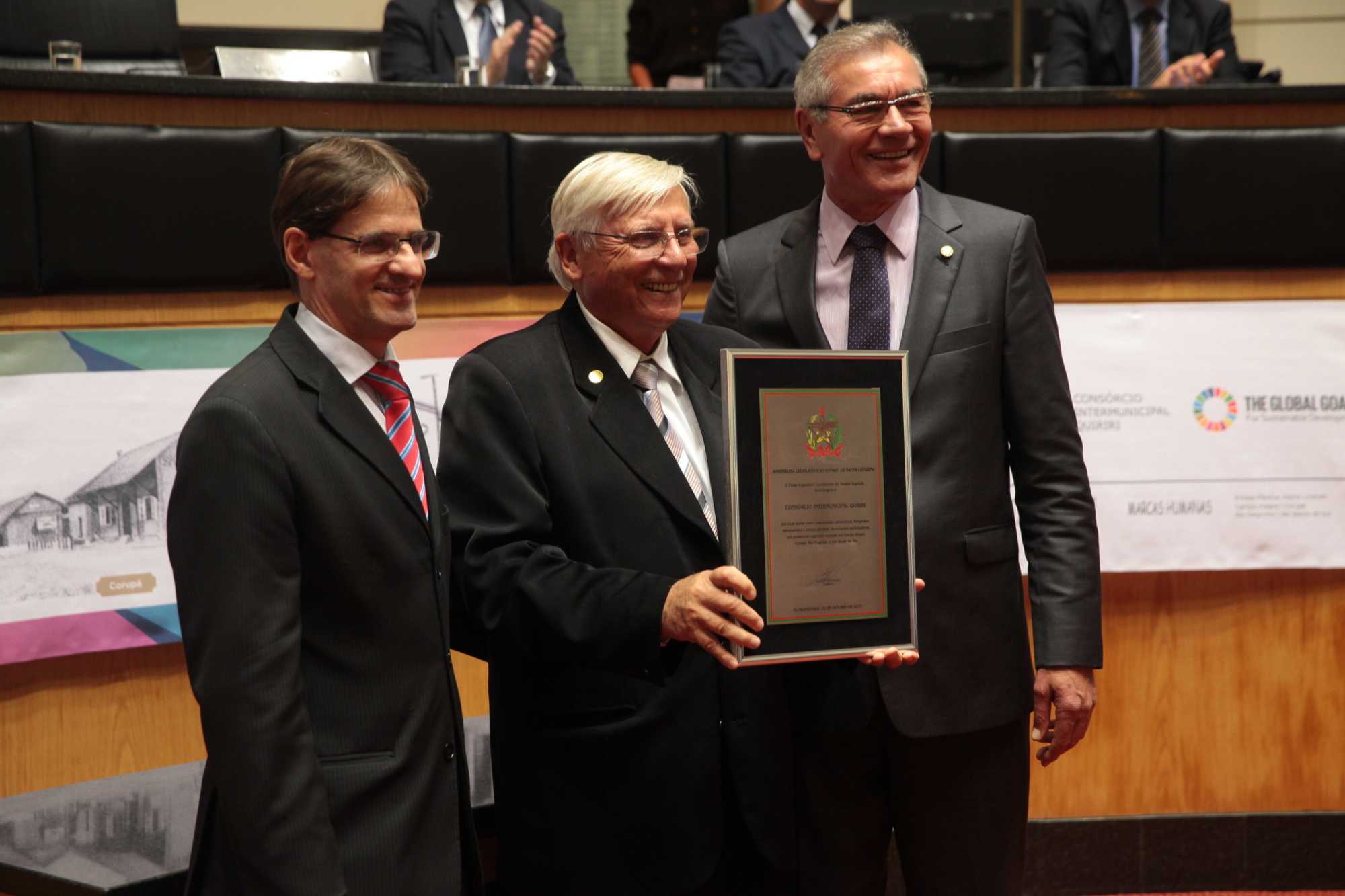 Deputados Neodi Saretta (à esquerda) e Silvio Dreveck entregam homenagem ao presidente do consórcio, Magno Bollmann (centro)